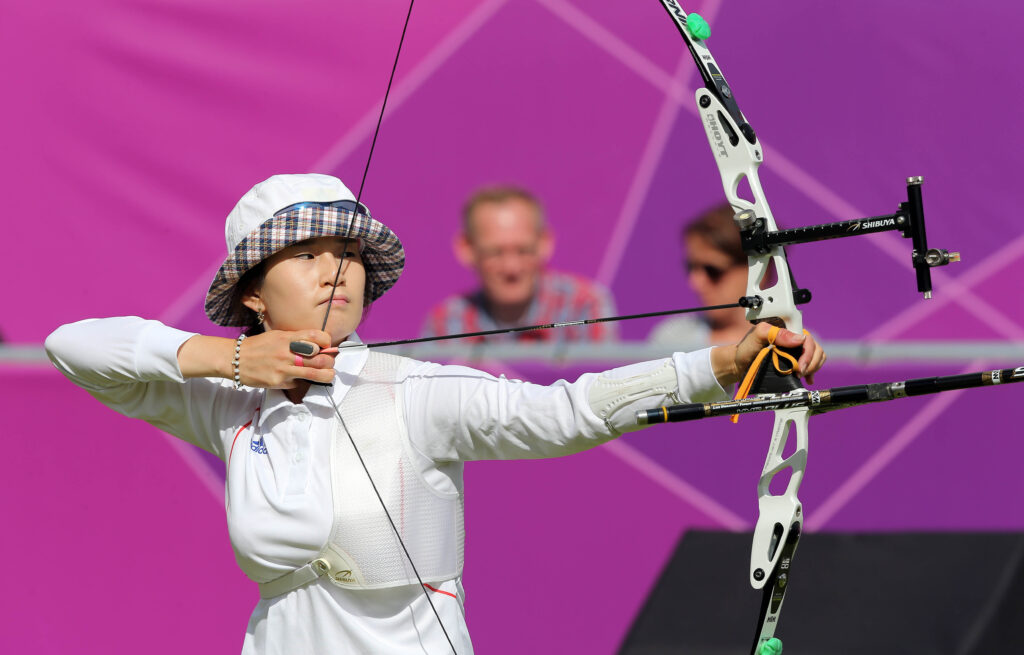 Olympic archer Ki Bo-bae aims a white Hoyt recurve bow against a purple geometric background. She wears a white polo shirt, a chest guard, and a plaid-rimmed bucket hat.